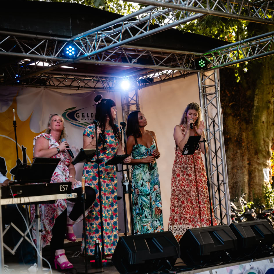 Vier Personen stehen auf einer B&uuml;hne unter einem Metallger&uuml;st mit Lichtern, alle tragen lange, bunte Kleider.