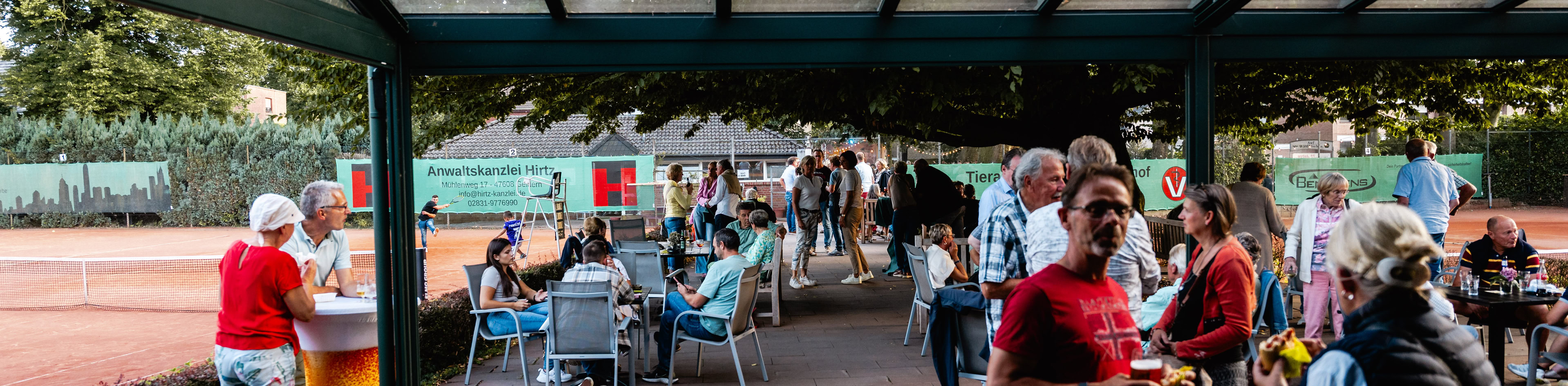 Menschen versammeln sich unter einem &uuml;berdachten Bereich neben einem Tennisplatz, einige sitzen an Tischen, w&auml;hrend andere stehen und sich unterhalten.