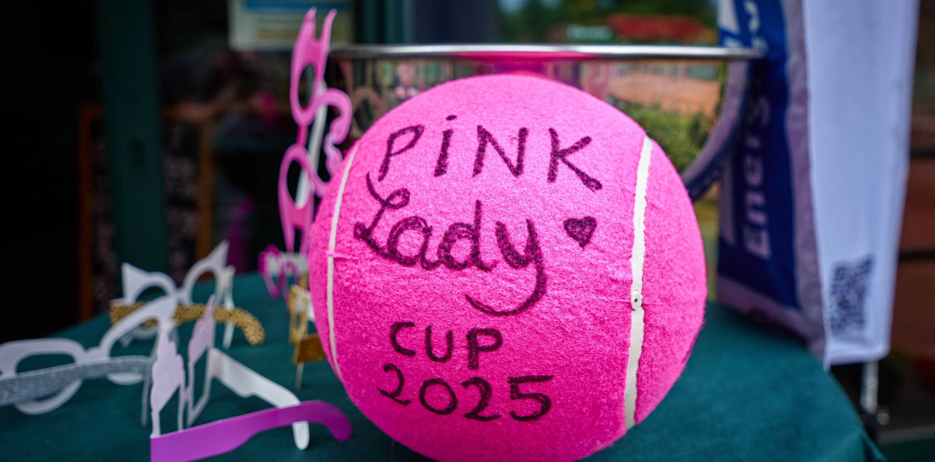 Ein pinker Tennisball mit der Aufschrift "Pink Lady Cup 2025" auf einem Tisch, umgeben von Dekorationen und einem Pokal im Hintergrund.