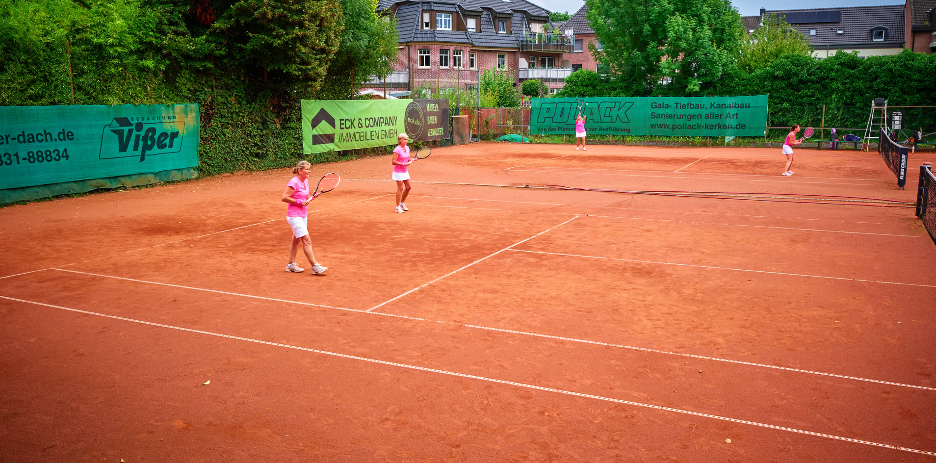 Tennisplatz mit vier Personen, die Tennis spielen; umgeben von B&auml;umen und Geb&auml;uden im Hintergrund.