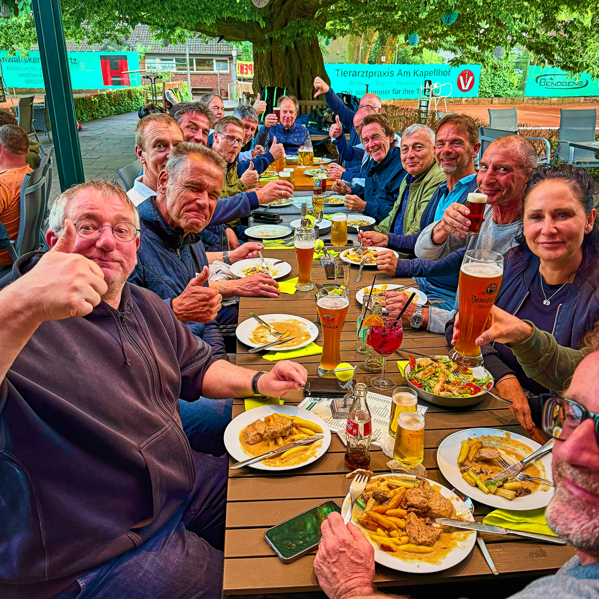 Eine Gruppe von Menschen sitzt an einem langen Tisch im Freien und isst und trinkt gemeinsam.
