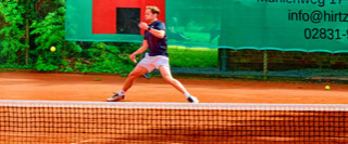 Eine Person spielt Tennis auf einem Sandplatz, umgeben von gr&uuml;ner Vegetation und einem gr&uuml;nen Banner im Hintergrund.