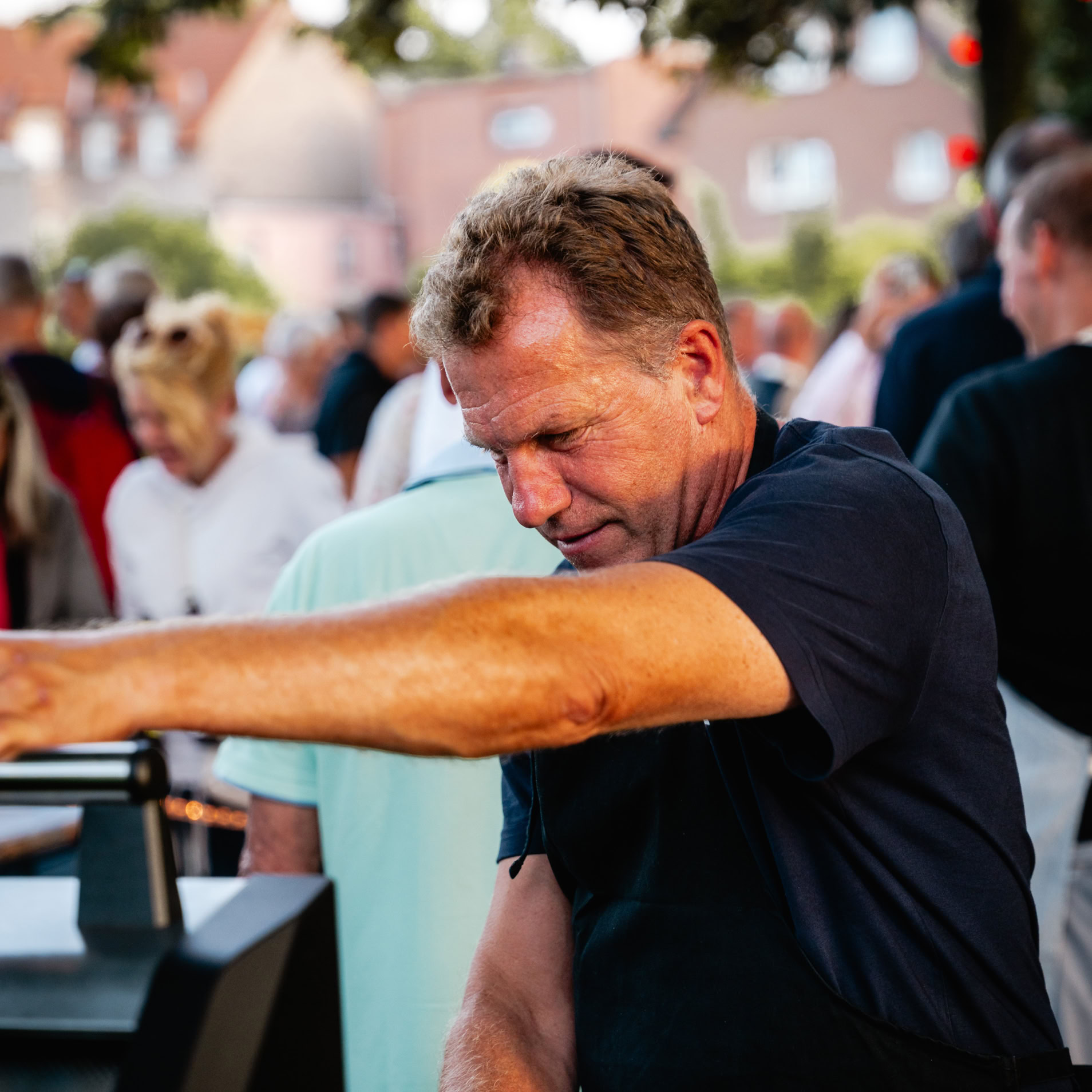 Mann in dunkelblauem T-Shirt bedient einen Grill bei einer Veranstaltung im Freien, umgeben von mehreren Personen.