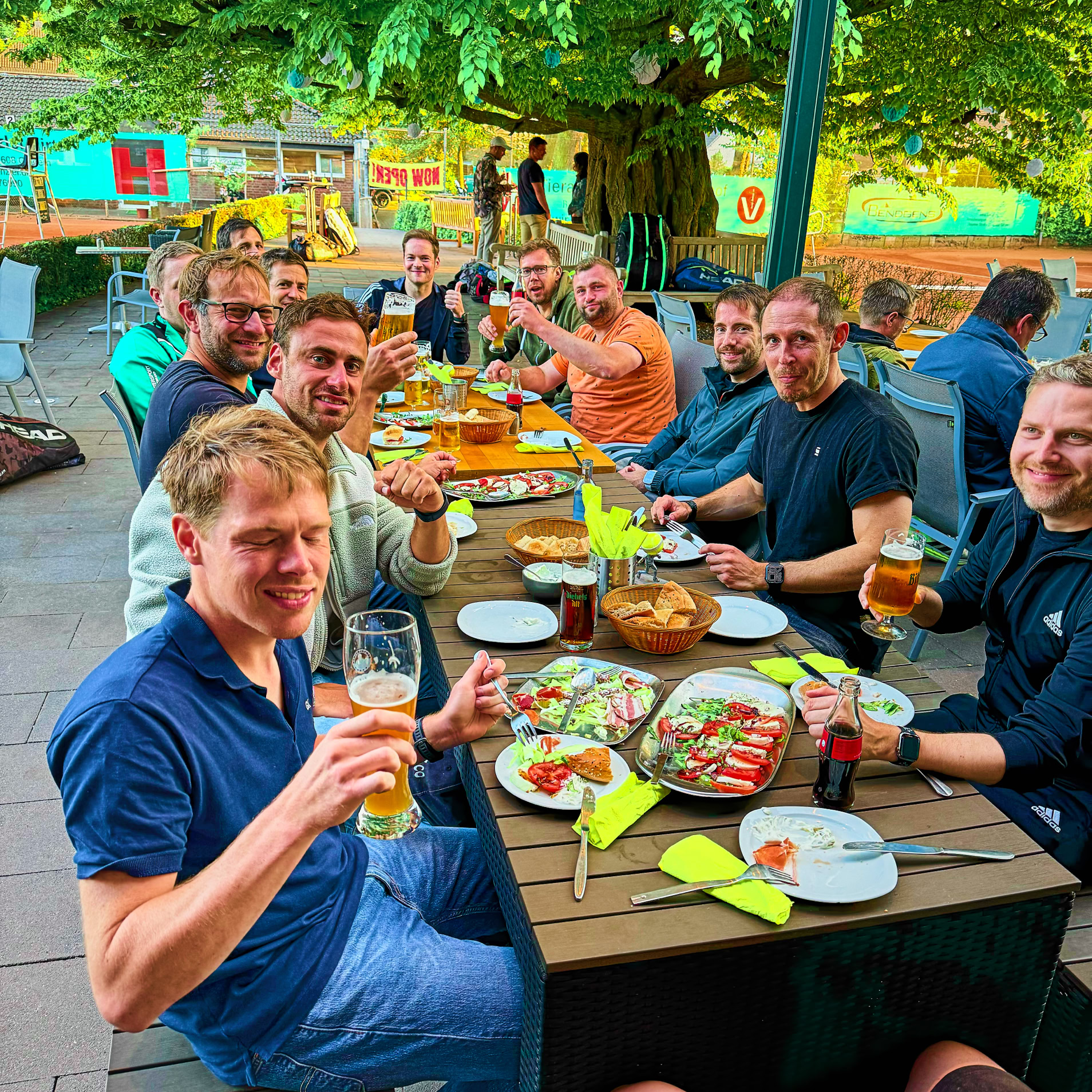 Gruppe von Menschen sitzt an einem langen Tisch im Freien, isst und trinkt.