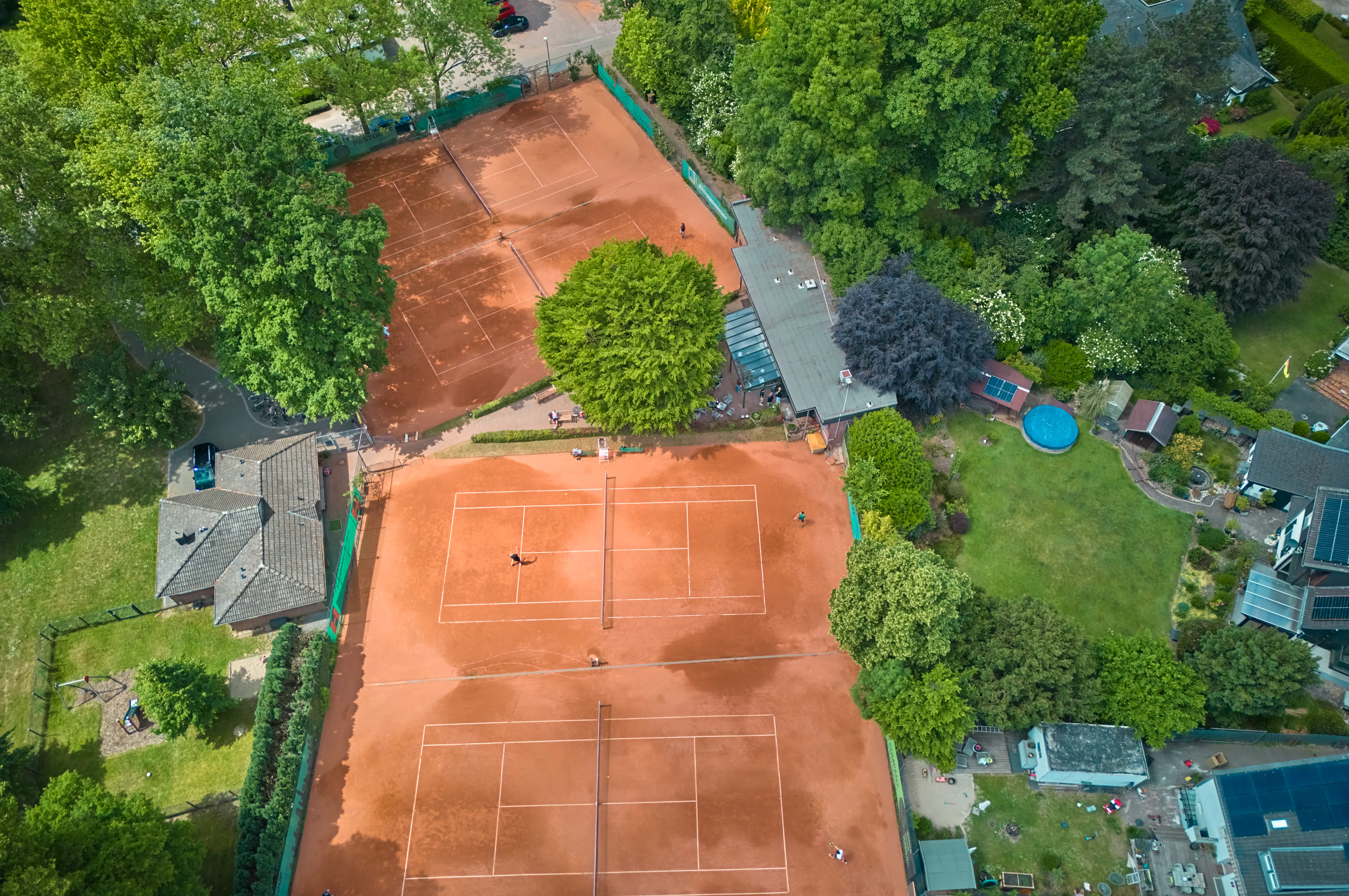 Luftaufnahme von zwei Sandtennispl&auml;tzen, umgeben von B&auml;umen und Geb&auml;uden.