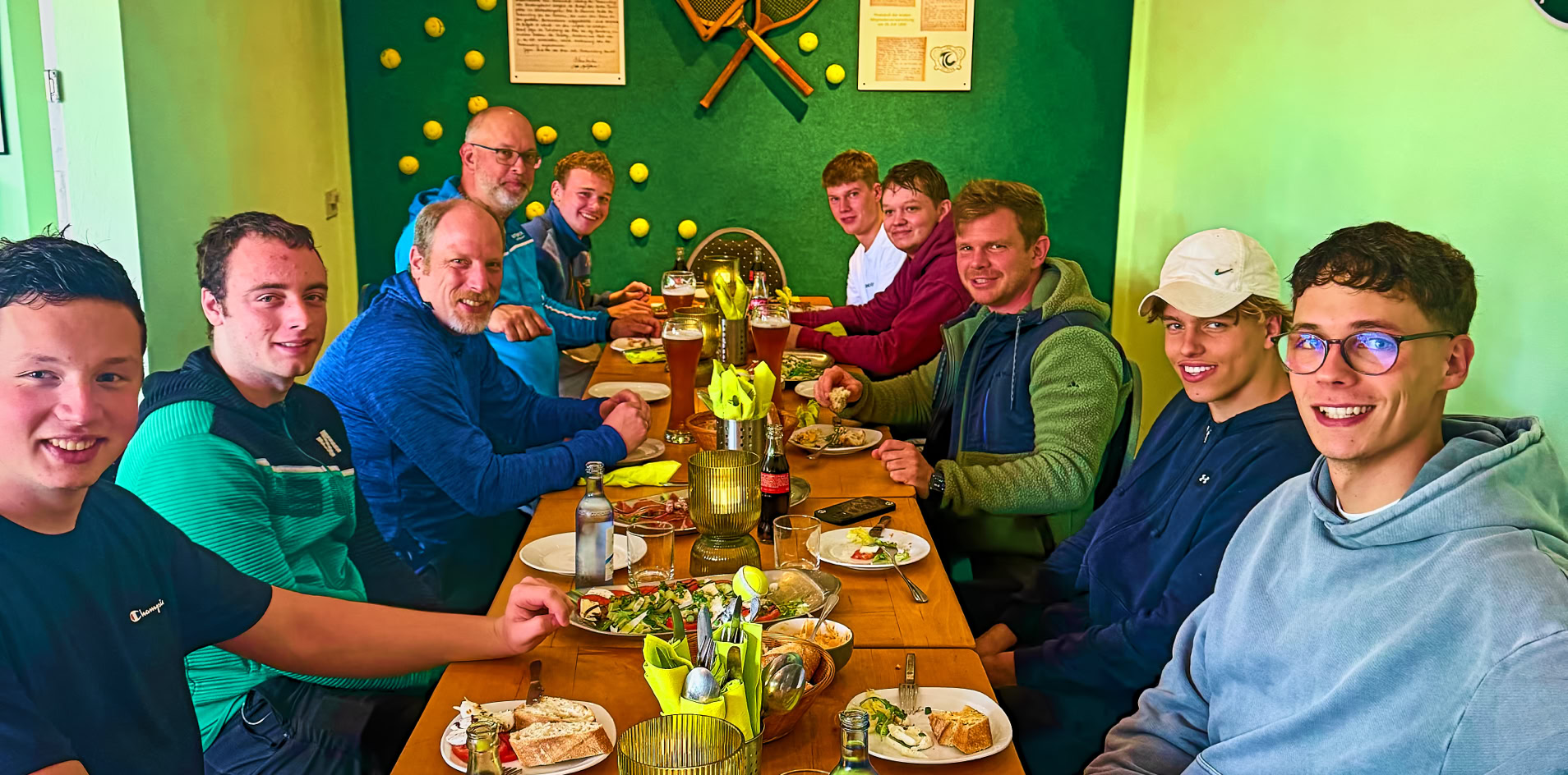 Eine Gruppe von Menschen sitzt an einem langen Tisch in einem Raum mit gr&uuml;nen W&auml;nden, dekoriert mit Tennisschl&auml;gern und B&auml;llen. Auf dem Tisch stehen Teller, Gl&auml;ser und Getr&auml;nke.
