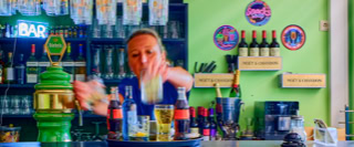 Eine Person steht hinter einer Bar und bereitet ein Getr&auml;nk zu. Im Hintergrund sind Flaschen und Gl&auml;ser auf Regalen zu sehen. Die W&auml;nde sind gr&uuml;n und es h&auml;ngen dekorative Schilder und Bilder.