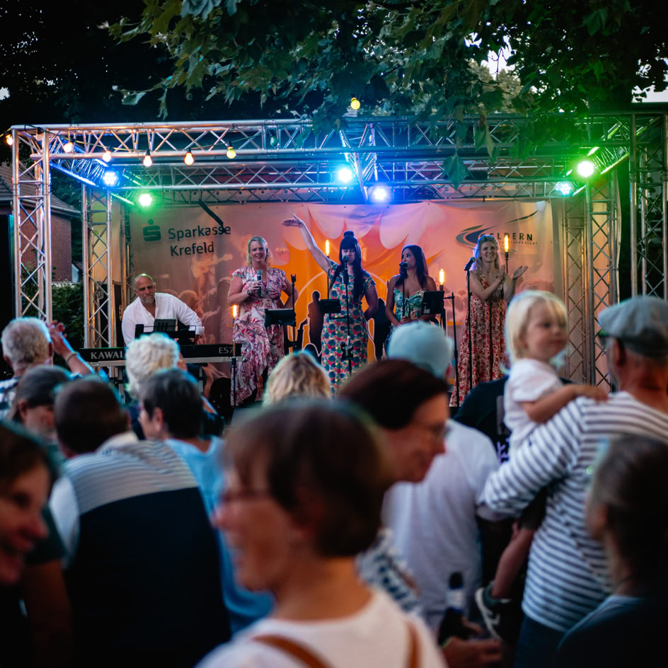 Menschenmenge vor einer B&uuml;hne mit bunten Lichtern, auf der mehrere Musiker auftreten.