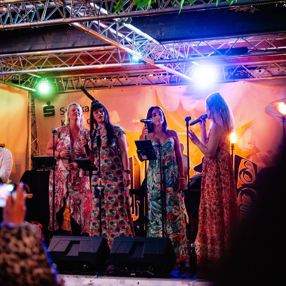 Vier Personen singen auf einer B&uuml;hne mit bunten Lichtern und tragen lange, gemusterte Kleider.