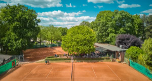 Tennisplatz mit rotem Sandboden, umgeben von B&auml;umen und einem Geb&auml;ude im Hintergrund unter blauem Himmel mit Wolken.