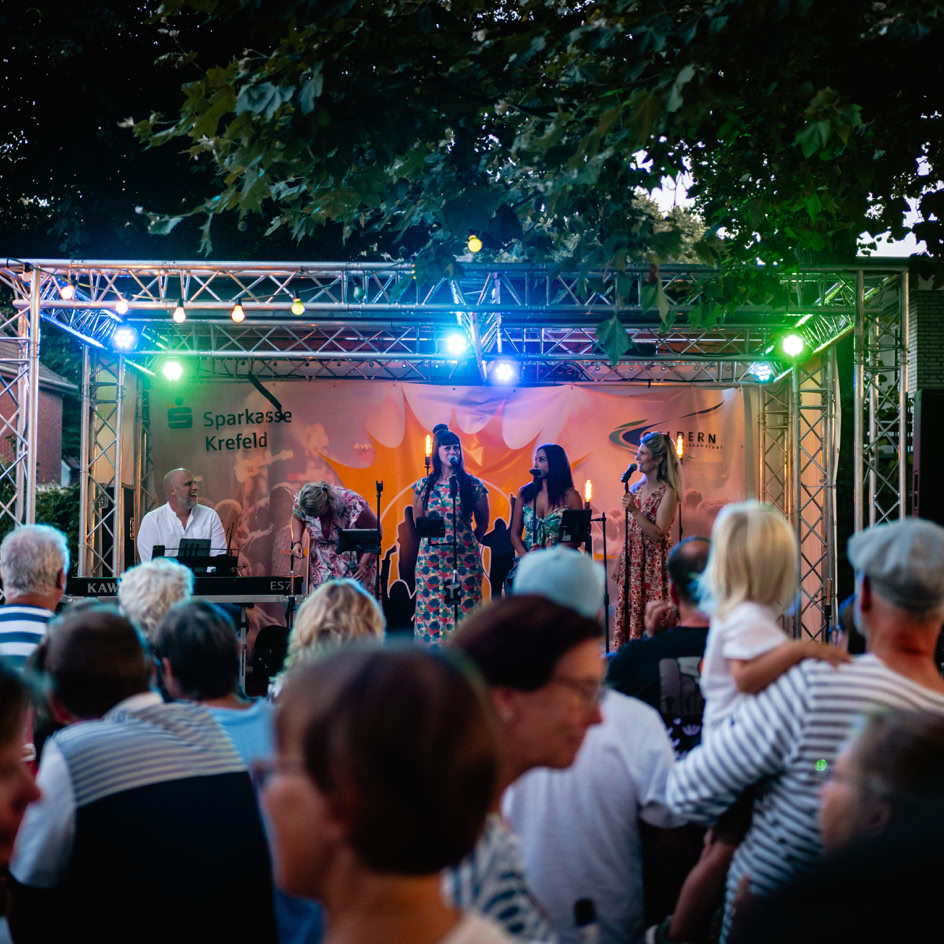 Eine Menschenmenge steht vor einer beleuchteten B&uuml;hne im Freien, auf der mehrere Personen auftreten.