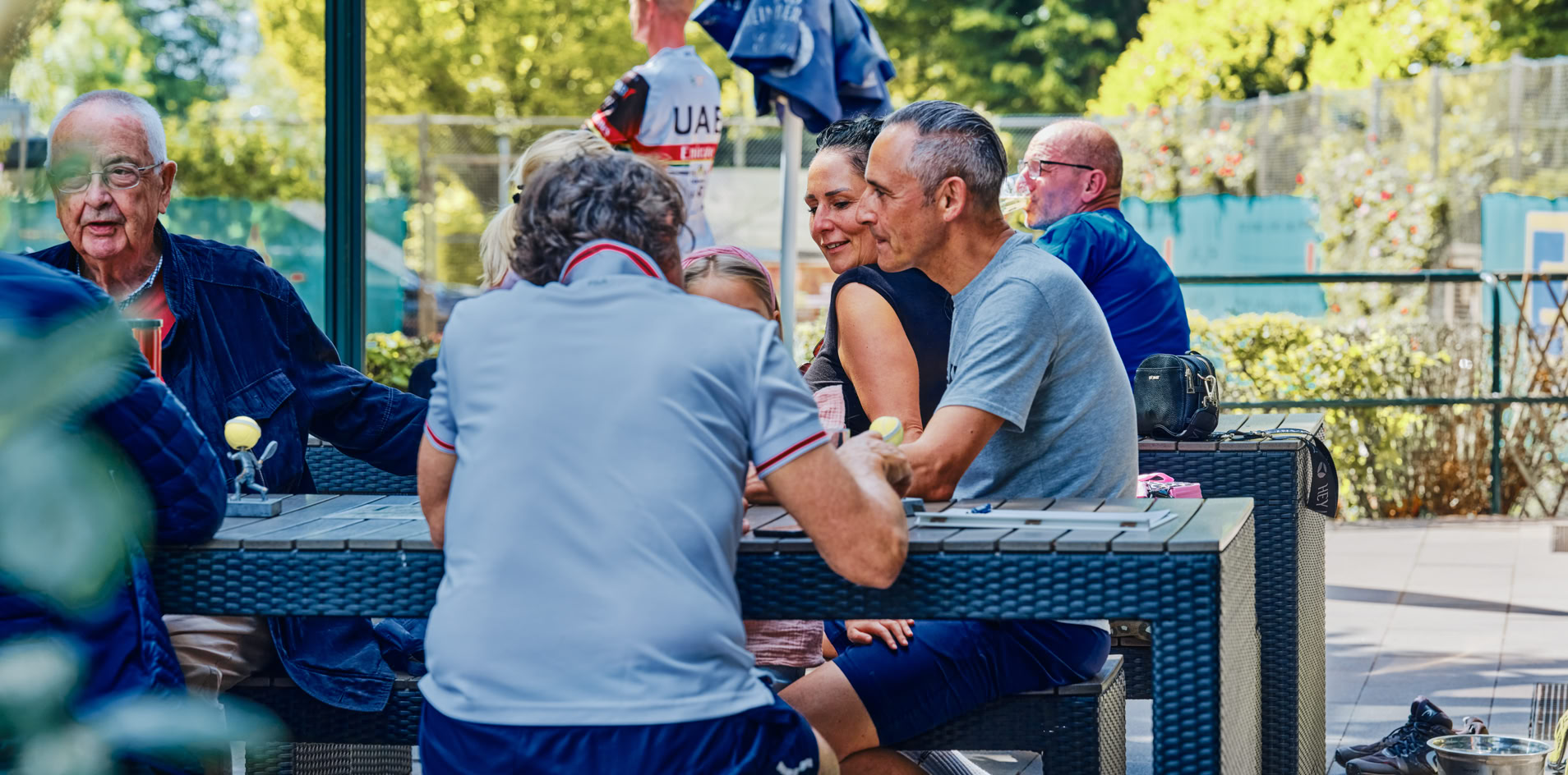 Eine Gruppe von Menschen sitzt an einem Tisch im Freien, umgeben von B&auml;umen und einem Zaun im Hintergrund.