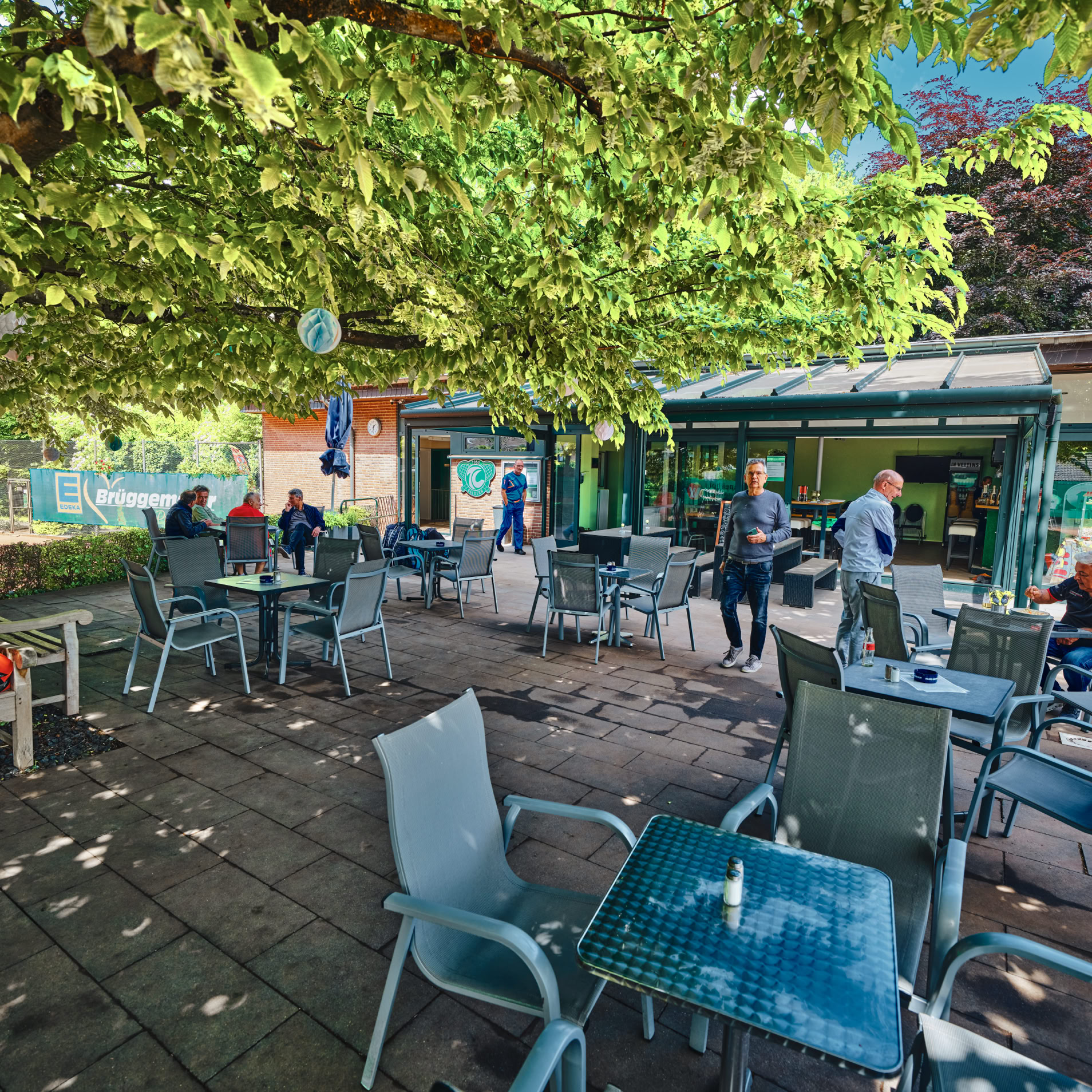 Au&szlig;enbereich eines Caf&eacute;s mit mehreren Tischen und St&uuml;hlen unter einem gro&szlig;en Baum, Menschen sitzen und stehen, Geb&auml;ude im Hintergrund.