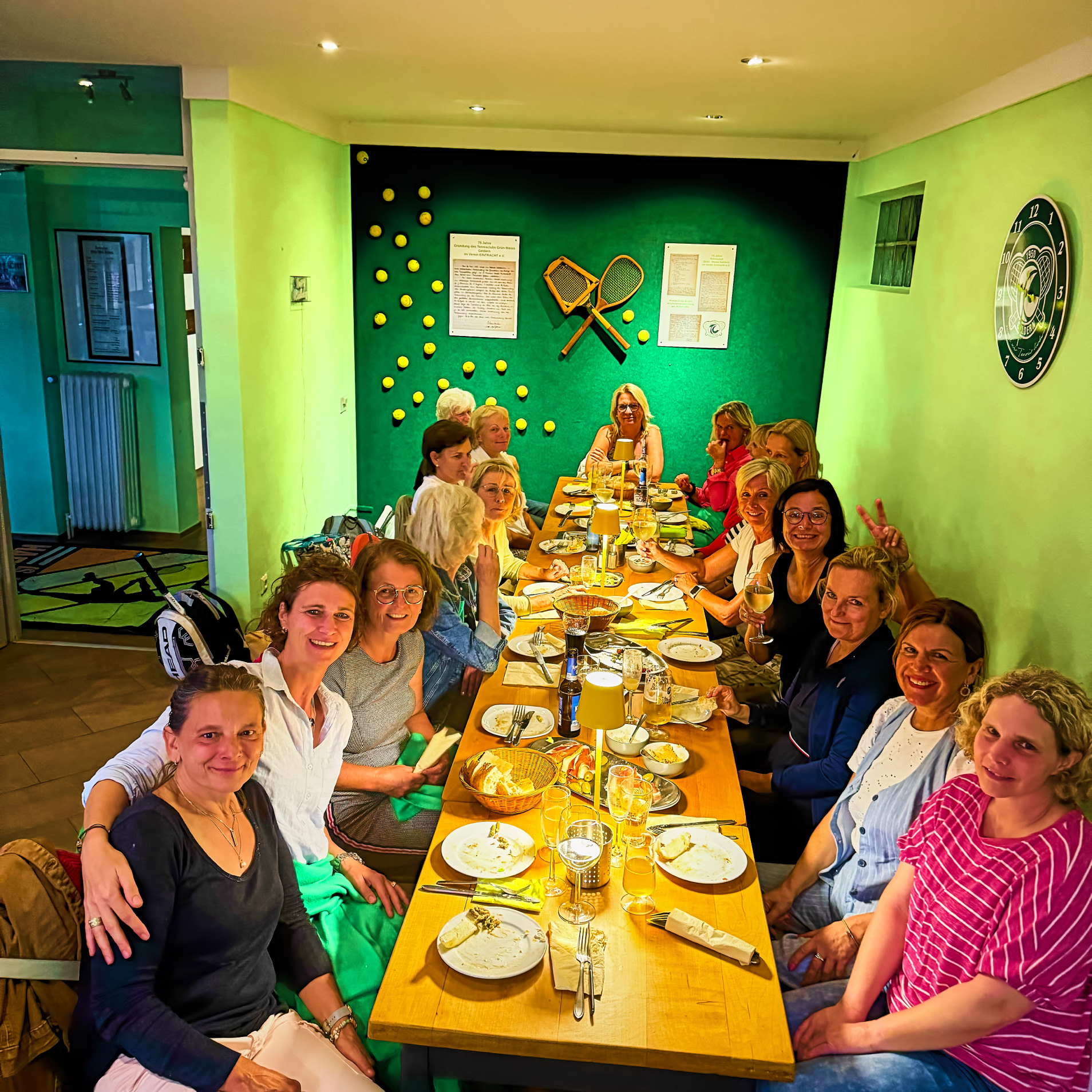 Eine Gruppe von Menschen sitzt an einem langen Tisch in einem Raum mit gr&uuml;nen W&auml;nden, auf dem Tisch stehen Teller und Gl&auml;ser.