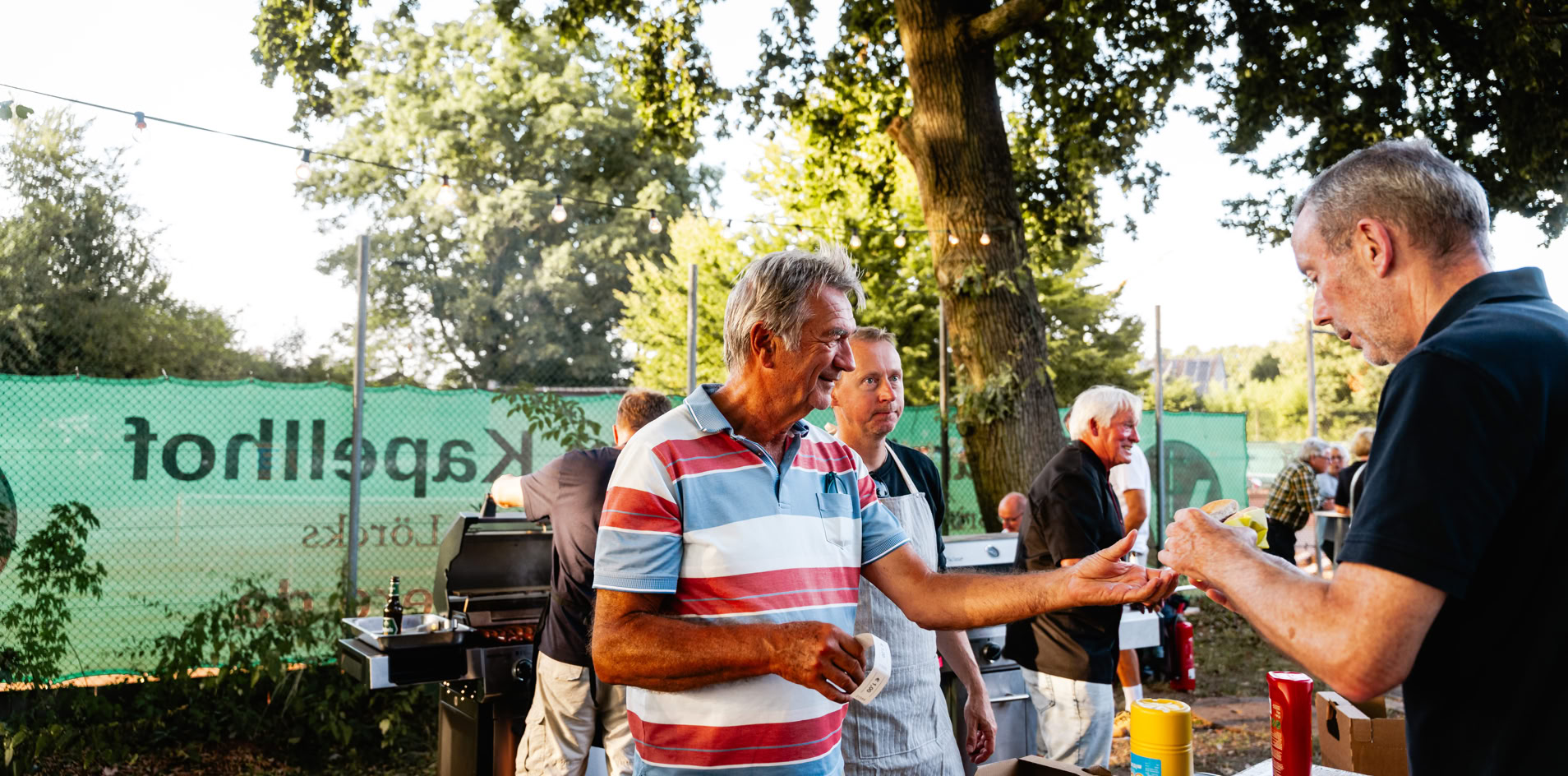 Mehrere Personen stehen im Freien an einem Tisch, umgeben von B&auml;umen und einem gr&uuml;nen Zaun. Ein Mann im gestreiften Hemd erh&auml;lt etwas von einem anderen Mann. Im Hintergrund ist ein Grill zu sehen.