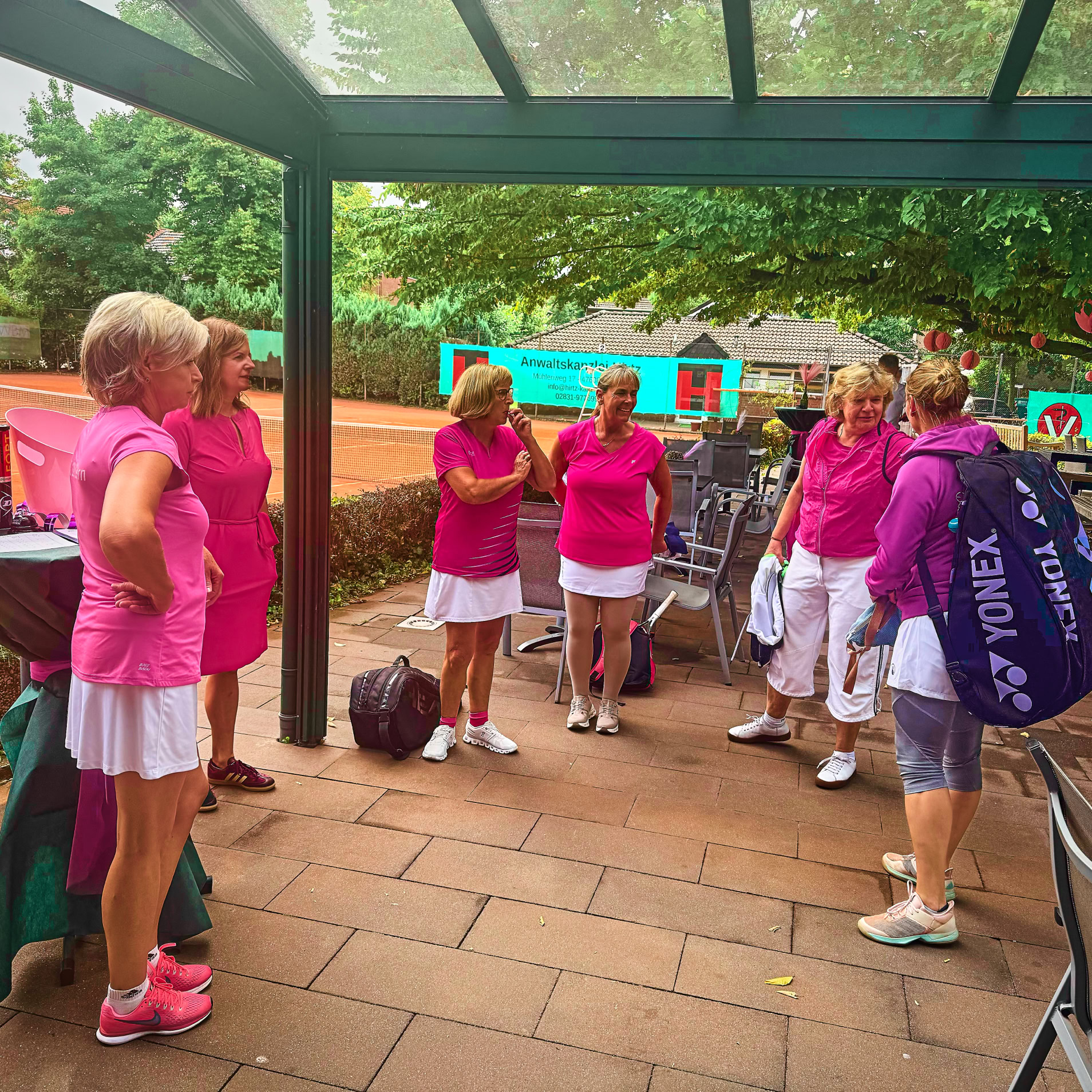 Eine Gruppe von Frauen in pinker Sportkleidung steht unter einem &uuml;berdachten Bereich neben einem Tennisplatz.
