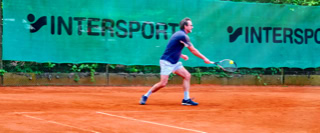 Ein Tennisspieler in blauer Kleidung auf einem Sandplatz, bereit, einen Ball zu schlagen, mit einem gr&uuml;nen Zaun im Hintergrund, auf dem "INTERSPORT" steht.