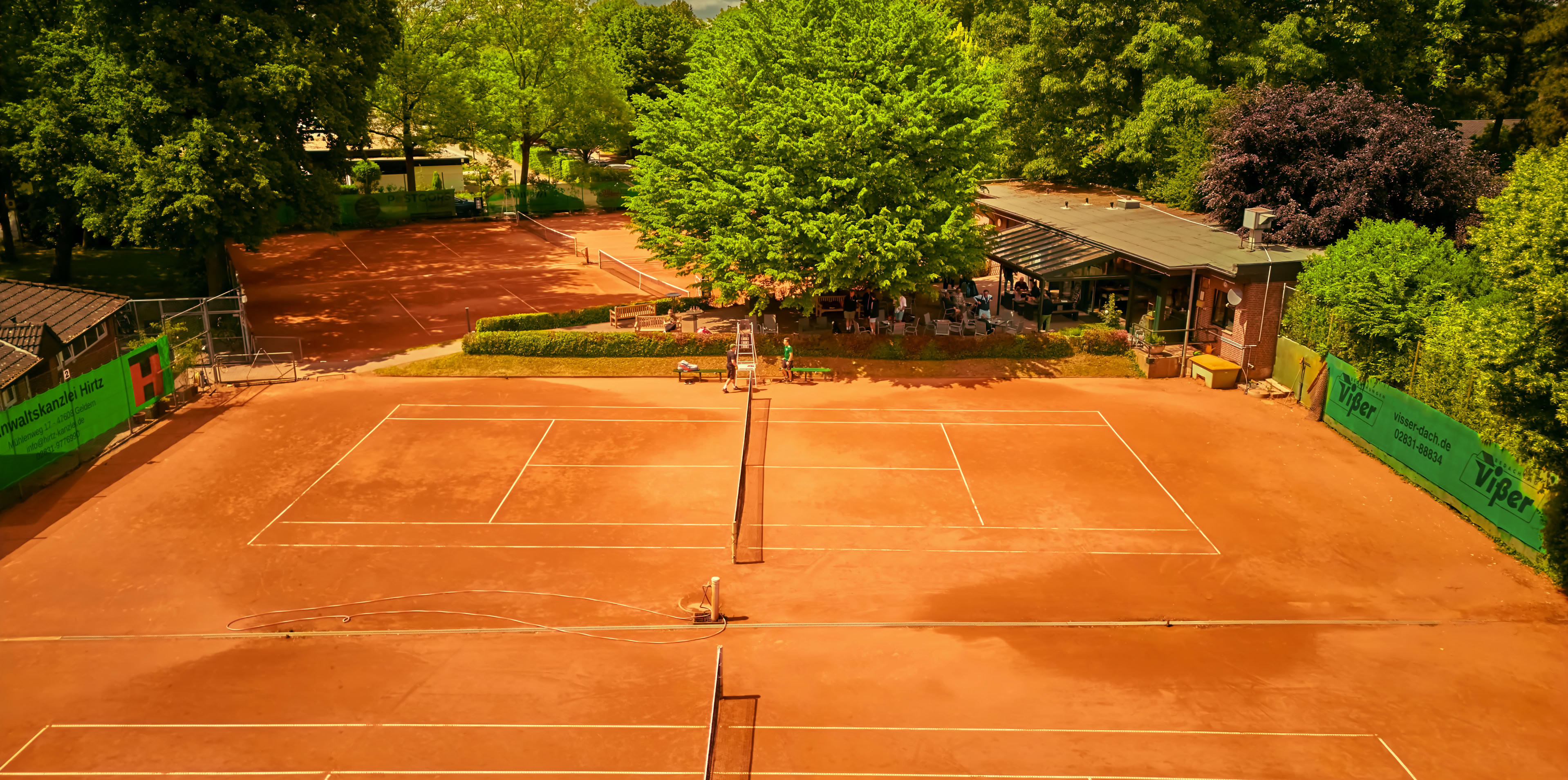 Zwei Sandtennispl&auml;tze umgeben von B&auml;umen und einem Geb&auml;ude im Hintergrund.