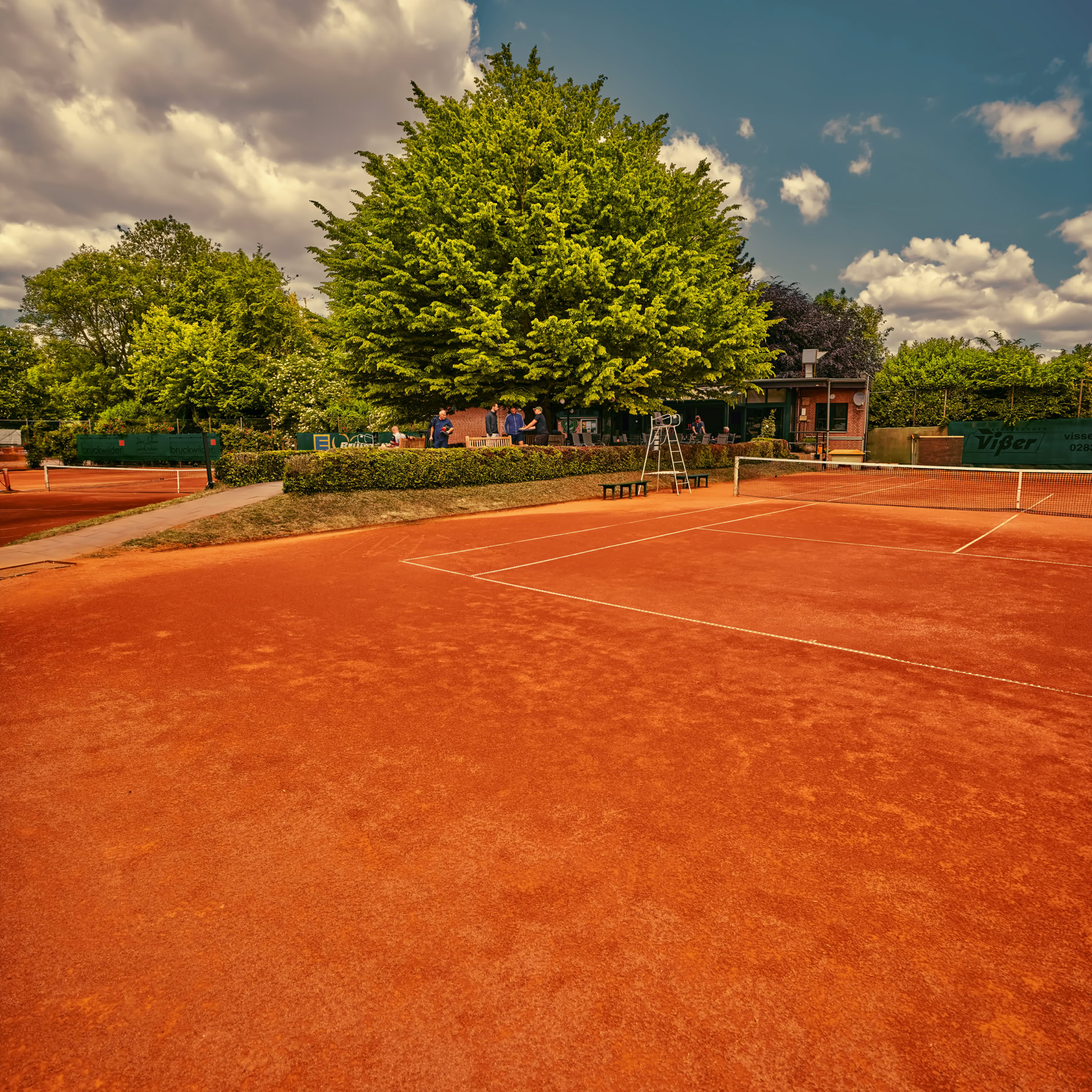 Ein roter Sandtennisplatz mit einem gro&szlig;en Baum im Hintergrund, umgeben von gr&uuml;ner Vegetation und einem bew&ouml;lkten Himmel.