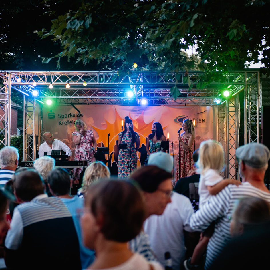 Eine Menschenmenge steht vor einer B&uuml;hne im Freien, auf der eine Band unter bunten Lichtern auftritt.